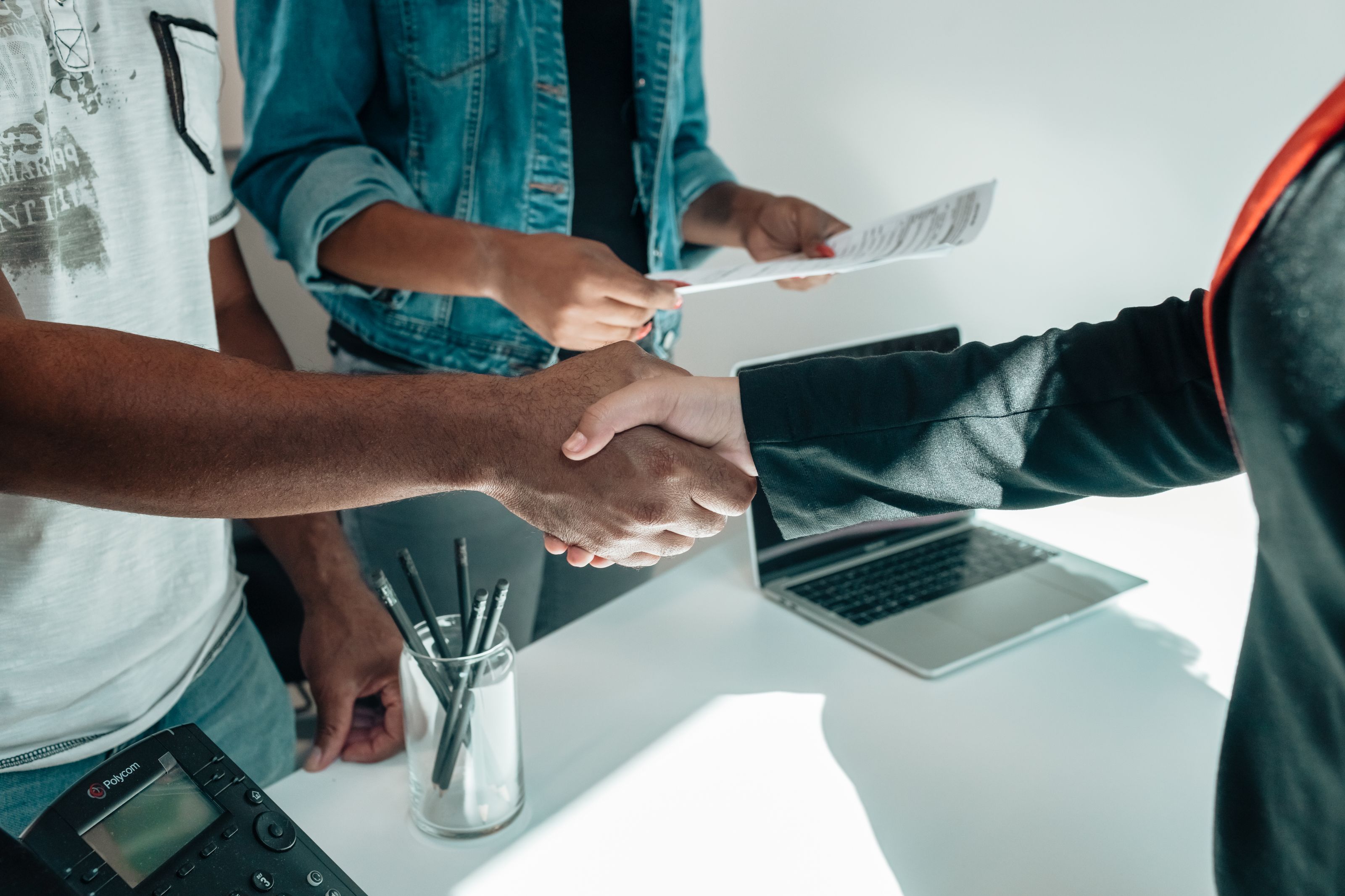 Two people shaking hands over a desk while a document is passed between them.